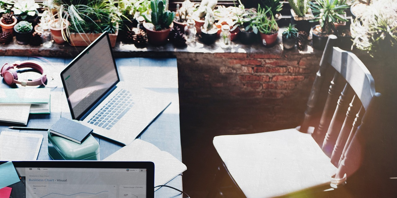 Overhead view of a laptop and notebooks on a desk near a window, with a rustic chair and various potted plants in the background.