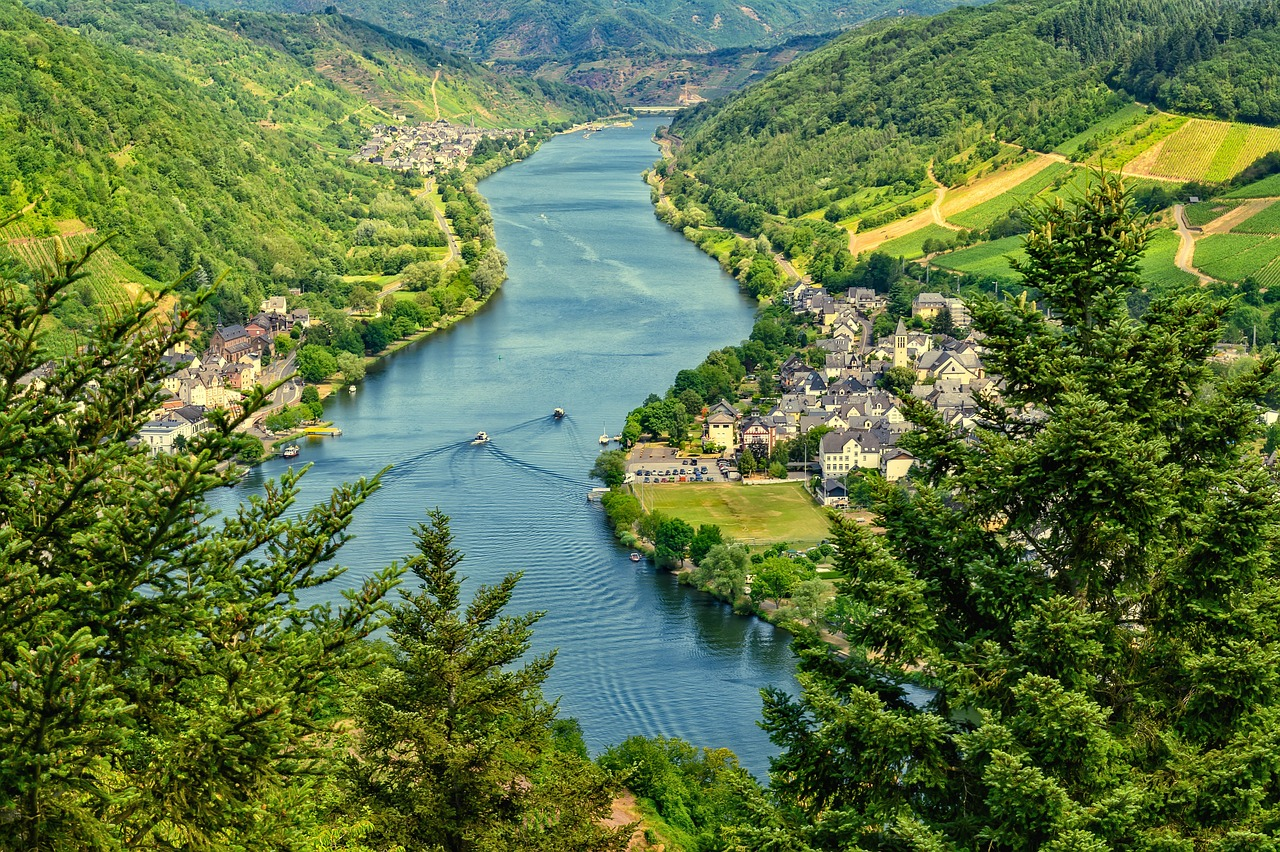 Aerial view of the Mosel River valley in Germany, winding between steep, green, forested hillsides with scattered vineyards and small clustered villages along the riverbanks.